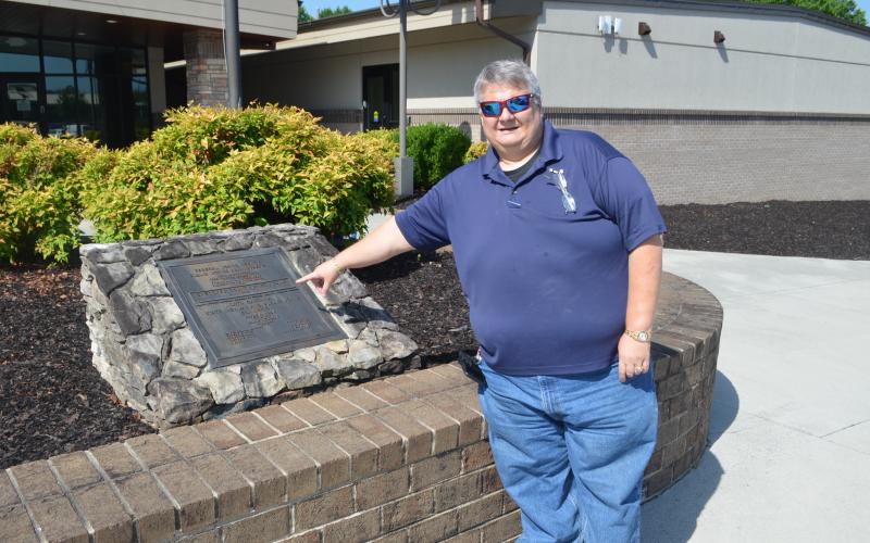 Abigail Hickman/Staff Correspondent Trevis Hicks points to the original plaque from the Cherokee County Prison Camp. Behind him is the original prison house that has been renovated into a classroom building.
