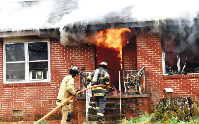 Becky Long • Clay County Progress. Firefighters move in closer to assess the situation amid smoldering eaves and flames still billowing from the interior front entrance way and window. The fire was contained but the house is most likely destroyed from the damages.