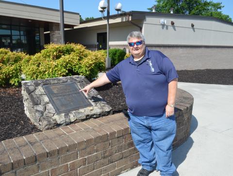 Abigail Hickman/Staff Correspondent Trevis Hicks points to the original plaque from the Cherokee County Prison Camp. Behind him is the original prison house that has been renovated into a classroom building.