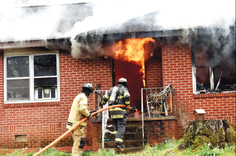Becky Long • Clay County Progress. Firefighters move in closer to assess the situation amid smoldering eaves and flames still billowing from the interior front entrance way and window. The fire was contained but the house is most likely destroyed from the damages.